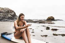 Mujer joven con tabla de surf sentada en la playa, usando smartphone — Stock Photo