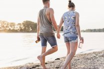 Молодая пара, гуляющая по солнечному берегу реки с бутылками — стоковое фото