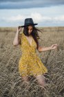 Young woman wearing summer dress with floral design and a hat dancing in corn field — Stock Photo
