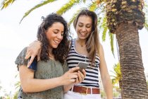 Two happy female friends looking at a smartphone under a palm tree — Stock Photo