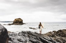 Jovem mulher carregando prancha de surf na praia rochosa no mar — Fotografia de Stock