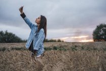 Jovem mulher tomando selfie com smartphone em um campo de milho ao pôr do sol — Fotografia de Stock