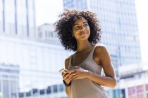 Smiling young woman with curly hair using cell phone in front of skyscrapers — Stock Photo