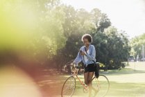 Young woman with cell phone pushing bicycle in park — Stock Photo