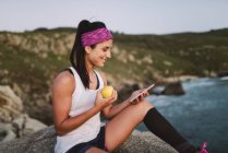 Спортивна жінка, що сидить на скелях увечері, тримає яблуко і за допомогою смартфона — стокове фото