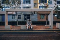 Homem do espaço sentado no banco na paragem de autocarro à noite com bebida — Fotografia de Stock