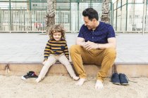 Spagna, Barcellona, padre e figlio seduti accanto alla spiaggia a piedi nudi — Foto stock