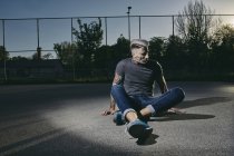 Jeune homme tatoué assis sur le terrain de basket au coucher du soleil — Photo de stock