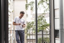 Hombre sonriente en pijama con taza de café de pie en el balcón - foto de stock