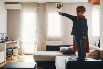 Kleiner Junge verkleidet als Superheld, der zu Hause auf einem Couchtisch steht und auf etwas zeigt — Stockfoto