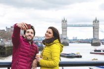 Royaume-Uni, Londres, jeune couple debout sur un pont au-dessus de la Tamise prenant selfie avec smartphone — Photo de stock
