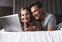 Sorridente coppia sdraiata sul letto a casa guardando il computer portatile — Foto stock