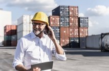 Uomo d'affari al porto di carico, con casco di sicurezza, con smartphone e tablet digitale — Foto stock