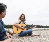 Счастливая женщина с мужчиной на берегу реки, играющим на гитаре — стоковое фото