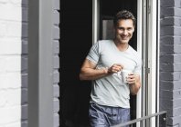 Ritratto di uomo sorridente in pigiama a casa con tazza di caffè alla porta del balcone — Foto stock