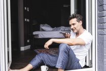 Uomo in pigiama a casa seduto alla finestra francese con smartphone — Foto stock