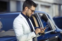 Успешный бизнесмен в городе, опираясь на автомобиль, используя смартфон — стоковое фото