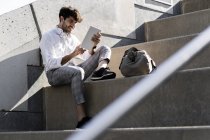 Улыбающийся молодой человек, сидящий на лестнице с помощью планшета — стоковое фото