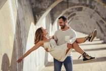 Spain, Andalusia, Malaga, happy man carrying girlfriend under an archway in the city — Stock Photo