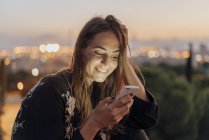 Іспанія, Барселона, Монжуїк, усміхнена молода жінка в сутінках за допомогою мобільного телефону — стокове фото