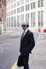 Retrato de elegante empresário sênior com óculos de sol e fones de ouvido em pé na calçada — Fotografia de Stock