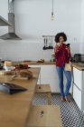 Женщина пьет белое вино на кухне, используя смартфон — стоковое фото