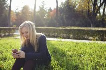 Donna bionda sorridente seduta su un prato nel parco cittadino autunnale, utilizzando lo smartphone — Foto stock