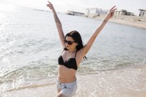 Feliz joven mujer caminando en la orilla del mar con los brazos levantados - foto de stock