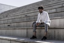 Stilvoller junger Mann mit Kopfhörern sitzt auf der Treppe im Freien — Stockfoto