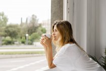 Молода жінка стоїть біля вікна і тримає чашку кави — стокове фото