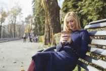 Portrait of blond woman sitting on bench in autumnal city park, using smartphone — Stock Photo
