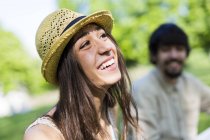 Portrait of happy young woman in a park with her boyfriend in the background — Stock Photo