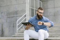 Ritratto di uomo d'affari hipster barbuto seduto sulle scale con tablet digitale e controllare l'ora — Foto stock