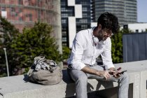Sorridente giovane uomo seduto su una panchina di cemento all'aperto utilizzando il telefono cellulare e sorridente — Foto stock