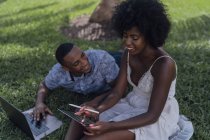 Happy young couple using tablet and laptop on lawn in a park — Stock Photo
