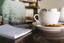 Чашка кави і блокнот на столі в кафе з жінкою на фоні — стокове фото
