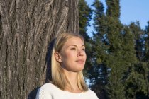 Портрет мечтающей блондинки, склоняющейся к стволу дерева и смотрящей на расстояние — стоковое фото