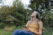 Femme avec téléphone portable assis dans le jardin et écouter de la musique avec écouteurs — Photo de stock