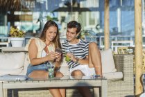Feliz pareja joven sentada en la terraza de un bar mirando el teléfono celular - foto de stock