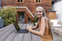 Portrait of smiling woman sitting on terrace of her house with laptop and glass of water — Stock Photo