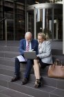 Homme d'affaires senior et femme d'affaires assis sur les escaliers avec ordinateur portable et graphiques — Photo de stock