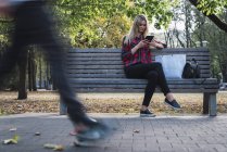 Young woman with shopping bag sitting on bench in autumn using mini tablet — Stock Photo