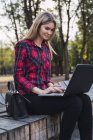 Giovane donna alla moda seduta sul banco all'aperto e che lavora sul computer portatile — Foto stock