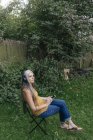Mujer con teléfono celular sentada en el jardín y escuchando música con auriculares - foto de stock