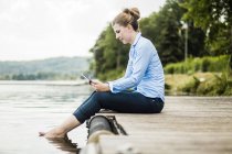Woman sitting on jetty at a lake and using tablet — Stock Photo