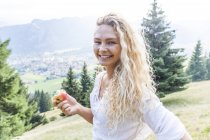 Alemania, Baviera, Oberammergau, retrato de una joven sonriente comiendo una manzana en el prado de la montaña - foto de stock