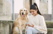 Молодая женщина со своей золотой собакой-ретривером на улице, используя мобильный телефон — стоковое фото