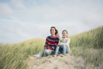 Mother and daughter sitting on a beach dune, girl pointing at distance, smiling — Stock Photo