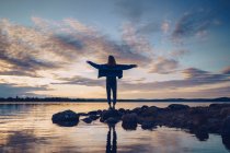 Молодая женщина, стоящая у озера Инари, с распростертыми руками, Финляндия — стоковое фото