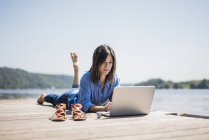 Femme mûre travaillant au bord d'un lac, utilisant un ordinateur portable sur une jetée — Photo de stock